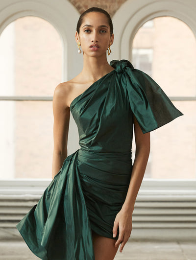 A model poses in a loft with exposed brick walls and large arched windows behind her, wearing a green bow-detailed cocktail dress with an asymmetrical hemline. 