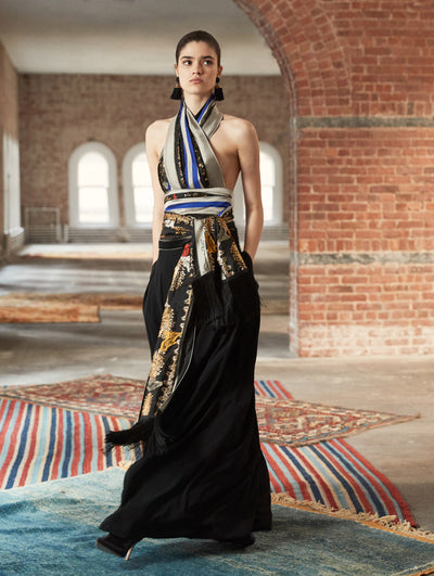 A model poses in a loft with exposed brick walls and large arched windows behind her, standing on a Persian rug. She wears a halter-neck blouse with a multi-colored patchwork print and floral-printed sash tied around her waist, paired with a black maxi skirt.
