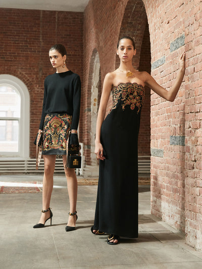 Two models pose in a loft with exposed brick walls, large arched windows and Persian rugs behind them. Holding a black cube-like handbag, the model on the left is wearing a black long-sleeve bow-detailed knit pullover and mini skirt with a red and gold floral tapestry design. The model on the right is wearing a black strapless column evening gown with a red and gold crystal-embroidered floral tapestry design concentrated at the bodice.
