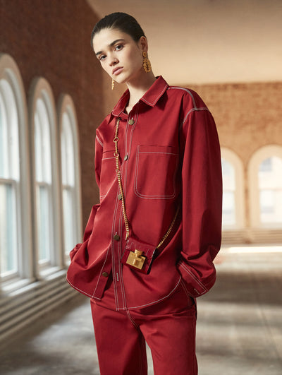 A model poses in a loft with exposed brick walls and large arched windows behind her, wearing a crossbody red micro push-lock bag, paired with a red long-sleeve button-up blouse and coordinating red trousers.