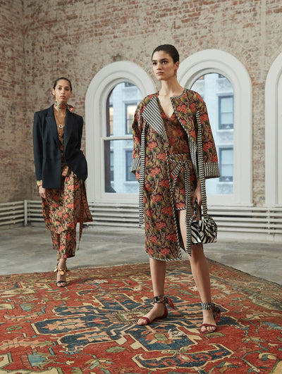 Two models pose in a loft with exposed brick walls and large arched windows behind them, standing on Persian rugs. The model on the left wears a black long-sleeve double-breasted blazer, layer over a red dress with a floral design. Holding a zebra print gardenia appliqué handbag, the model on the right wears a an open-front multi-colored partwork quilt coat, layered over a mini dress of the same pattern.