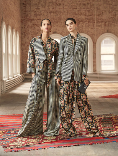 Two models pose in a loft with exposed brick walls and large arched windows behind them, standing on Persian rugs. The model on the left wears an open-front patchwork cropped quilt coat and silk wide leg pants printed with a geometric circle motif. Holding a black push-lock clutch, the model on the right wears a long-sleeve double-breasted blazer printed with a geometric circle design, paired with wide-leg silk trousers printed with a floral tapestry design.