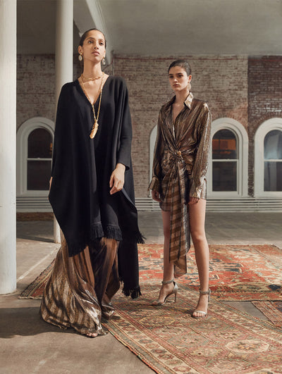 Two models pose in a loft with exposed brick walls and large arched windows behind them, standing on Persian rugs. The model on the left wears a black oversized long-sleeve knit tunic with fringe trim, layered over metallic stripped wide-leg pants. The model on the right wears a metallic striped mini dress with long-sleeve and drape detailing at the waist.