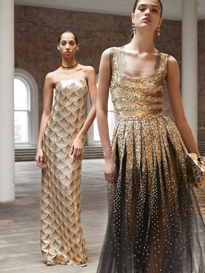 Two models pose in a loft with exposed brick walls and large arched windows behind them, standing on Persian rugs. The model on the left wears a strapless column gown with all-over scalloped bugle bead embroidery. Holding a gold bead-embroidered clutch, the model on the right wears a sleeveless evening gown with gold sequin embroidery concentrated at the bodice fading into a pleated scatter toward the skirt.