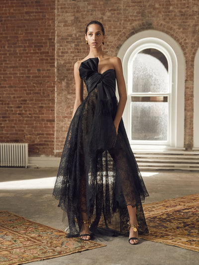 A model poses in a loft with exposed brick walls and large arched windows behind her, standing on a Persian rug. She wears a black evening gown with a bow-detailed bodice and semi-sheer lace skirt with an asymmetrical hemline.