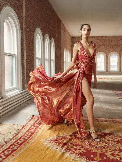 A model poses in a loft with exposed brick walls and large arched windows behind her, standing on a Persian rug. She wears a red sleeveless V-neck evening down with drape detailing and a gold metallic sheen.