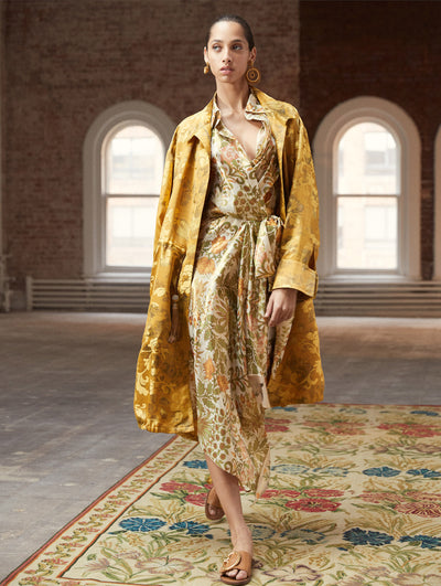 A model poses in a loft with exposed brick walls and large arched windows behind her, standing on a Persian rug. She wears a gold open-front long-sleeve coat with a floral design, layered over a silk V-neck dress featuring a gold floral design.
