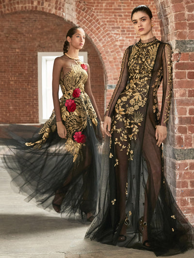 Two models pose in a loft with exposed brick walls and large arched windows behind them. The model on the left wears a black long-sleeve illusion-neck evening gown with a gold threadwork embroidered tapestry design and red florals. The model on the left wears a black long-sleeve semi-sheer evening gown with a gold threadwork embroidered tapestry design.