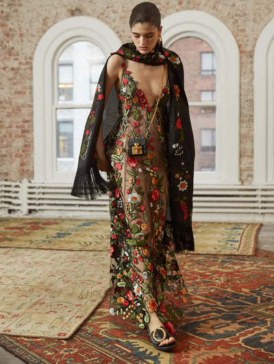 A model poses in a loft with exposed brick walls and large arched windows behind her, standing on Persian rugs. She wears a crossbody black micro push-lock bag and a black floral-embroidered scarf with fringe trim, layered over a black semi-sheer sleeveless cocktail dress with multi-colored threadwork embroidered florals.