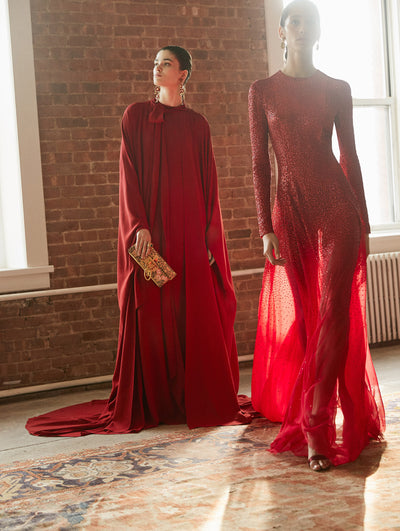 Two models pose in a loft with exposed brick walls and large arched windows behind them, standing on a Persian rug. Holding a floral-printed push-lock clutch, the model on the left wears a red long-sleeve bow-detail caftan. The model on the right wears a red long-sleeve semi-sheer evening gown with sequin embroidery concentrated on the bodice that fades to a delicate scatter towards the skirt.