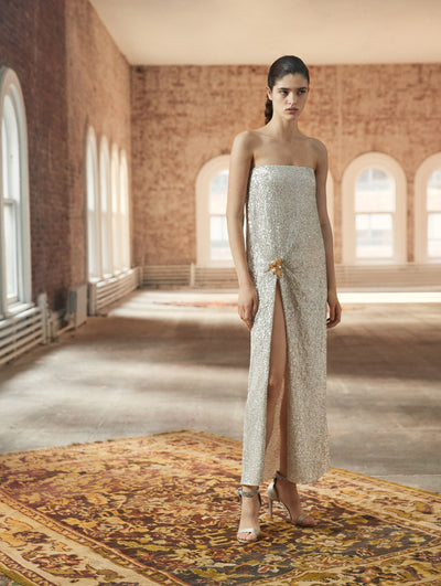 A model poses in a loft with exposed brick walls and large arched windows behind her, standing on a Persian rug. She wears a dazzling white strapless column gown with a front slit and gold leaf detail at the waist.