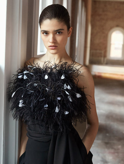 A model poses in a loft with exposed brick walls and large arched windows behind her. She wears a black jumpsuit with a black and white feather bodice.