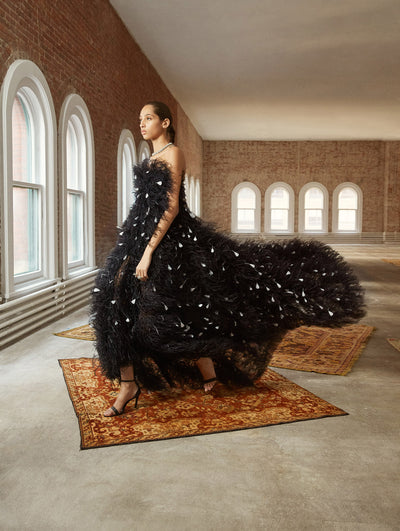 A model poses in a loft with exposed brick walls and large arched windows behind her, standing on a Persian rug. She wears a strapless high-low gown made entirely of black and white feathers.