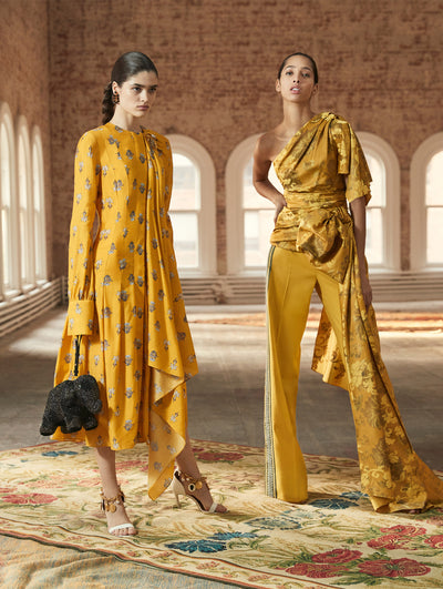 Two models pose in a loft with exposed brick walls and large arched windows behind them, standing on a Persian rug. Holding a black elephant-shaped handbag, the model on the left wears a yellow long-sleeve dress with a floral design and drape detailing. The model on the right wears a gold one-shoulder blouse with a floral design, paired with gold trousers.