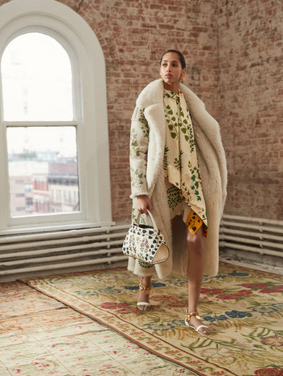 A model poses in a loft with exposed brick walls and large arched windows behind her, standing on a Persian rug. Holding a floral-printed top handle bag, she wears a cream long-sleeve open-front fur coat with a floral-printed exterior, layered over a cream dress printed with the same floral-printed motif.
