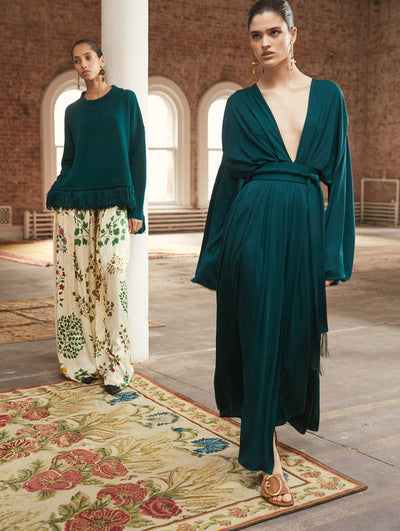 Two models pose in a loft with exposed brick walls and large arched windows behind them, standing on a Persian rug. The model on the left wears a green long-sleeve knit pullover with fringe trim, paired with cream floral-printed trousers. The model on the right wears a green V-neck long-sleeve jersey dress.