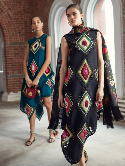 Two models pose in a loft with exposed brick walls and large arched windows behind them. Holding a red cube-like handbag, the model on the left wears a green sleeveless knit dress with an asymmetrical hemline and multi-colored crochet patchwork design. The model on the right wears a black sleeveless knit dress with an asymmetrical hemline and multi-colored crochet patchwork design, paired with a black floral-printed scarf with fringe trim.