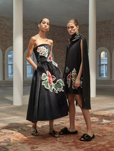 Two models pose in a loft with exposed brick walls and large arched windows behind them, standing on a Persian rug. The model on the left wears a black strapless cocktail dress with threadwork embroidered florals. The model on the right wears a black sleeveless shift dress with threadwork embroidered florals, paired with a black scarf with fringe trim.