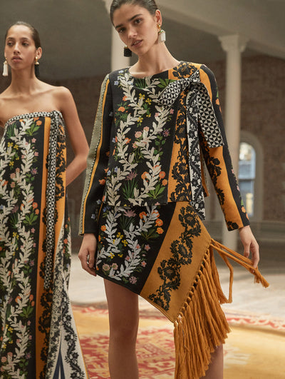 Two models pose in a loft with exposed brick walls and large arched windows behind them, standing on a Persian rug. The model on the left wears a strapless bow-detailed evening gown with a multi-colored patchwork print. The model on the right wears a long-sleeve bow-detailed knit jacket with a multi-colored patchwork print, paired with a knit mini skirt with fringe trim and the same multi-colored patchwork print.