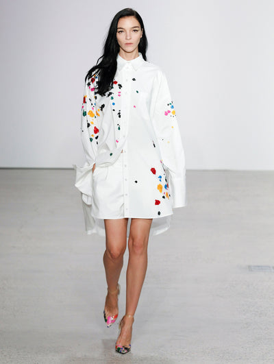The model walks the runway in a white button-up long-sleeve blouse embroidered with multi-colored sequin paint splashes and white shorts.