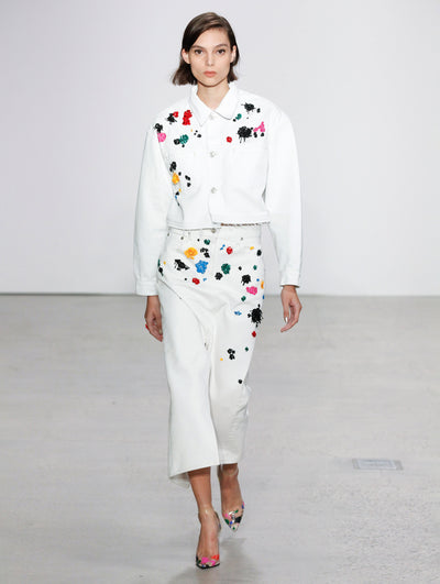 The model walks the runway in a white button-up long-sleeve jacket embroidered with multi-colored sequin paint splashes and a white coordinating skirt embroidered with multi-colored sequin paint splashes.