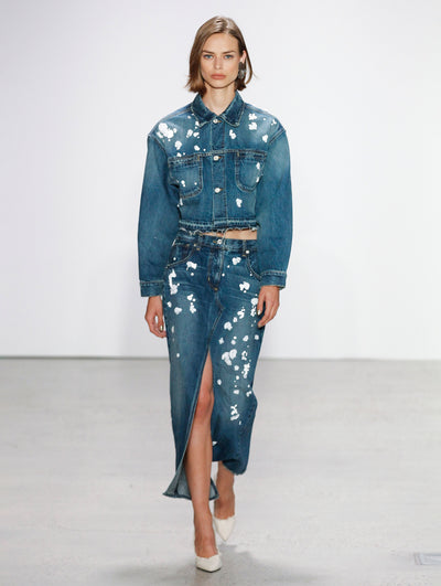 The model walks the runway in a denim button-up jacket printed with white paint splashes and a denim coordinating skirt printed with white paint splashes.