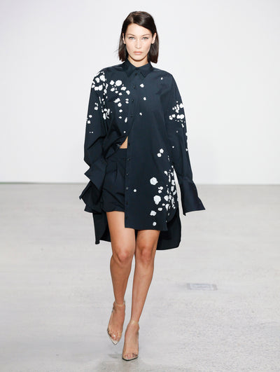 The model walks the runway in a navy button-up long-sleeve blouse printed with white paint splashes and navy shorts.