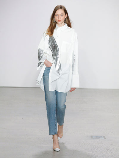 The model walks the runway in a white button-up long-sleeve blouse printed with a silver brushstroke across the chest and denim ankle-length pants.