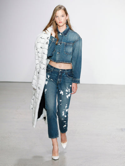 The model walks the runway in a button-up long-sleeve denim jacket and ankle-length denim pants printed with white paint splashes, while holding a white fur jacket embroidered with the Oscar de la Renta logo in black.
