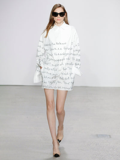 Wearing black sunglasses, the model walks the runway in a white button-up long-sleeve shirt dress that is printed with black cursive text. 