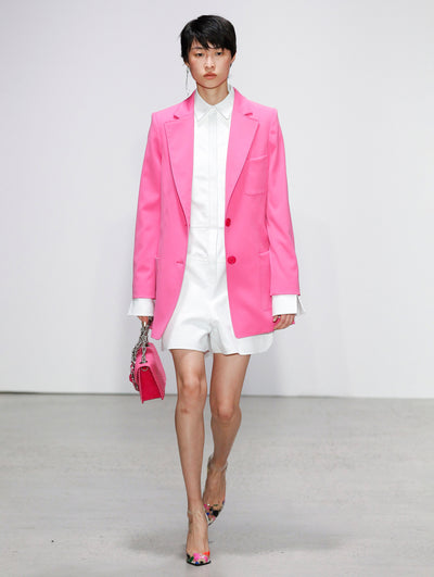 Holding a pink gardenia appliqué handbag, the model walks the runway in a pink double-breasted long sleeve blazer and white button-up long-sleeve romper.