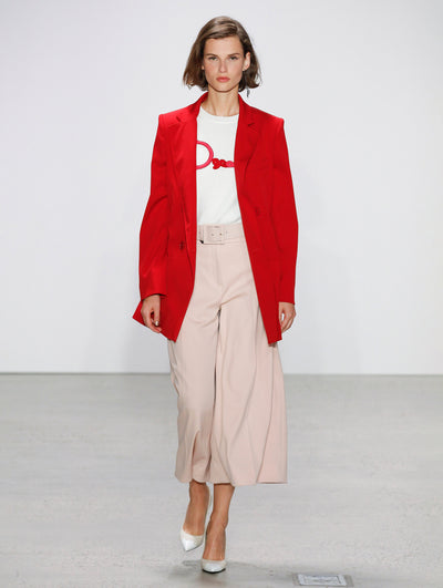 The model walks the runway in a red double-breasted long-sleeve blazer, logo printed white tee and belted blush trousers.