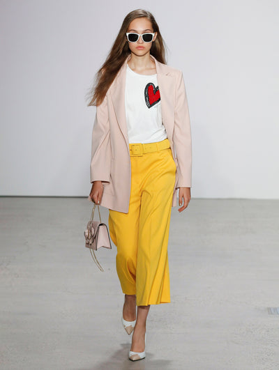 Wearing white sunglasses and holding a blush gardenia appliqué handbag, the model walks the runway in a blush double-breasted long-sleeve blazer, heart-printed white tee and belted yellow trousers.