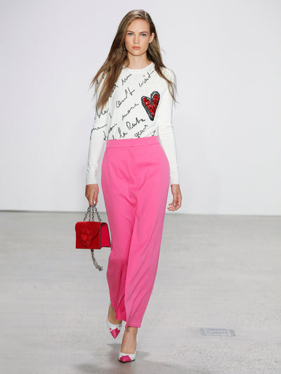 Holding a red gardenia appliqué handbag, the model walks the runway in logo and heart-printed long-sleeve white top and pink high-waisted trousers.
