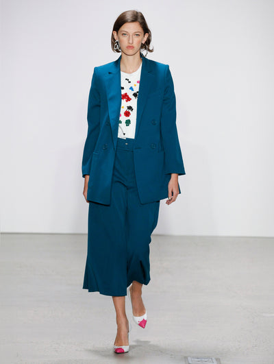 The model walks the runway in a blue double-breasted long sleeve blazer, belted coordinating blue trousers and a white tee embroidered with multi-colored sequin paint splashes.