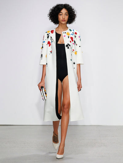 Holding a white clutch embroidered with multi-colored sequin paint splashes, the model walks the runway in a white open-front 3/4 sleeve jacket embroidered with multi-colored sequin paint splashes and a black bodysuit.