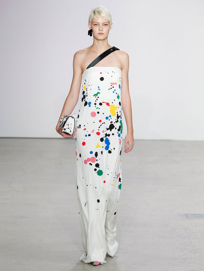 Holding a white gardenia appliqué handbag printed with multi-colored paint splashes, the model walks the runway in a white one-shoulder gown printed with multi-colored paint splashes.
