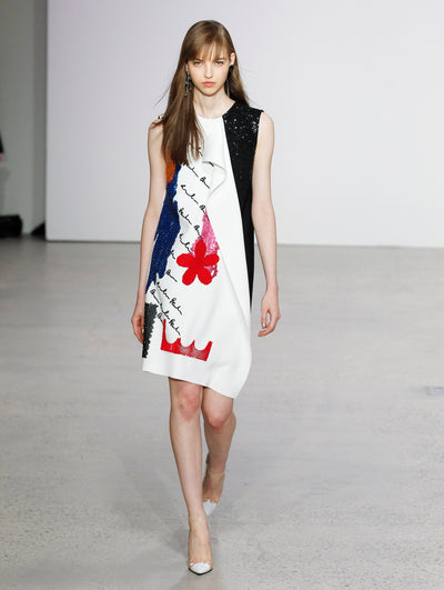 The model walks the runway in a white sleeveless cocktail dress embroidered with colorful flowers and organic forms and features black cursive text.