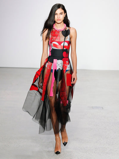 The model walks the runway in a semi-sheer black midi dress printed with red logo-filled brushstrokes, solid pink brushstrokes and black florals, layered over a sleeveless black, white, red and blue bodysuit, paired with a pink rope-like necklace.