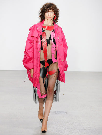 The model walks the runway in a pink oversized trench coat layered over a semi-sheer black midi dress printed with red and pink logo-filled brushstrokes, solid red and pink brushstrokes, and black florals.