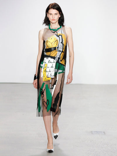 The model walks the runway in a semi-sheer black midi dress printed with white logo-filled brushstrokes, solid black and green brushstrokes, and yellow florals, layered over a sleeveless black, white, yellow and green bodysuit, paired with a green rope-like necklace.