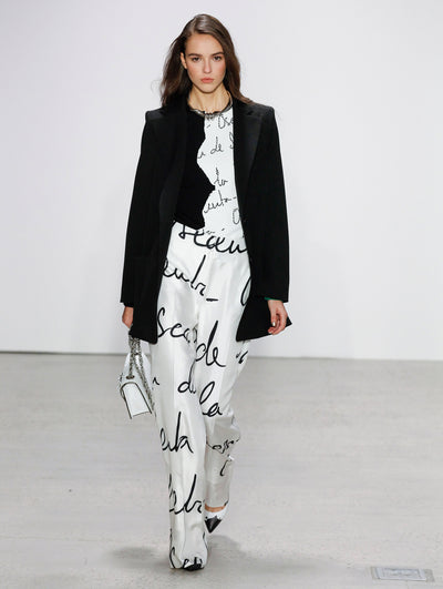 Holding a white gardenia appliqué handbag, the model walks the runway in black long-sleeve open-front coat, layered over a logo-printed white blouse and coordinating logo-printed white trousers, paired with a silver rope-like necklace.