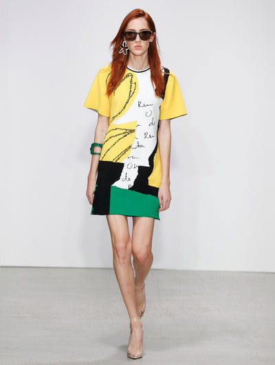 Wearing black sunglasses, the model walks the runway in a short-sleeve shift mini dress printed with white logo-filled brushstrokes, solid yellow, black and green brushstrokes, and black florals, paired with a green rope-like bangle necklace.