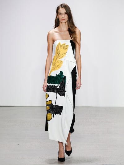 The model walks the runway in a strapless white draped cocktail dress embroidered with green and black sequin brushstrokes and yellow florals.