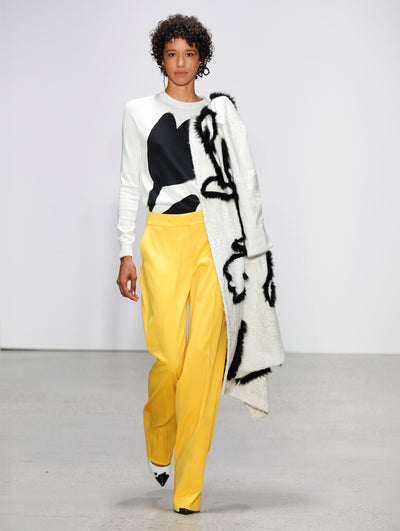 Holding a black and white long-sleeve fur coat, the model walks the runway in a floral-printed long-sleeve top and yellow trousers.