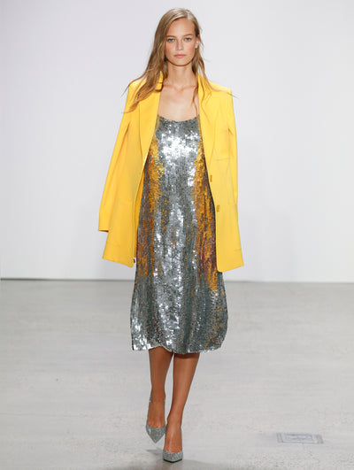 The model walks the runway in a yellow double-breasted long-sleeve blazer, layered over a silver all-over sequin cocktail dress.