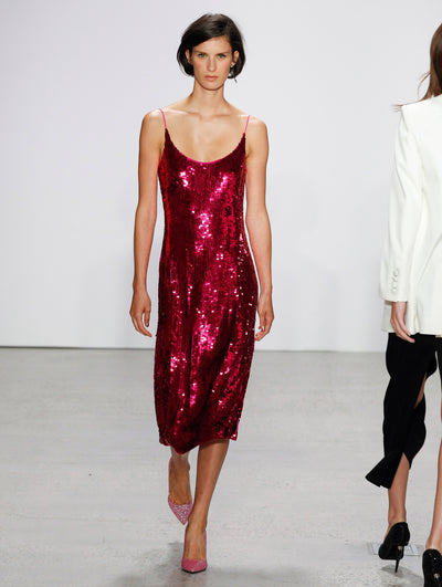 A model walks the runway in a pink all-over sequin sleeveless cocktail dress, as a second model in a white long-sleeve blazer and black ankle-length trousers passes by in the opposite direction, her back turned.