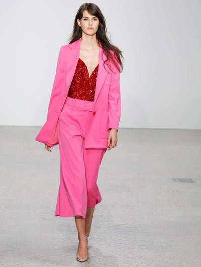 The model walks the runway in a pink double-breasted blazer, layered over coordinating belted pink trousers and an all-over sequin red blouse.