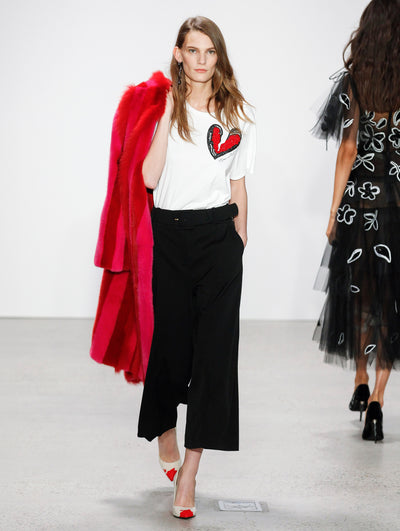 The model walks the runway in a heart-printed white tee and black belted trousers, holding a red fur coat, as a second model in a semi-sheer tulle cocktail dress with white floral embroidery passes by in the opposite direction, her back to the audience.