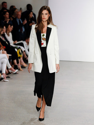 A model walks the runway in a white single-breasted long-sleeve blazer with black lapels, layered over a multi-colored sequin embroidered white top and black maxi skirt, with the audience visible in the background.